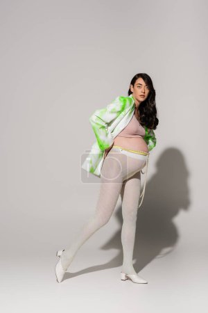Foto de Longitud completa de morena y encantadora mujer embarazada en leggings, top de la cosecha, chaqueta verde y blanco posando sobre fondo gris, concepto de moda de maternidad, expectativa - Imagen libre de derechos