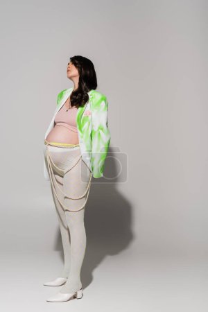 full length of fashionable pregnant woman in tights, crop top, green and white jacket and beads belt standing on grey background, maternity fashion concept, expectation 