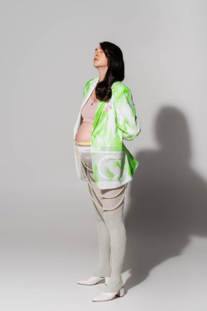 volle Länge der Schwangeren mit geschlossenen Augen, die in grün-weißer Jacke, bauchfreiem Oberteil, Perlengürtel und Leggings auf grauem Hintergrund steht, Umstandskonzept, Erwartung