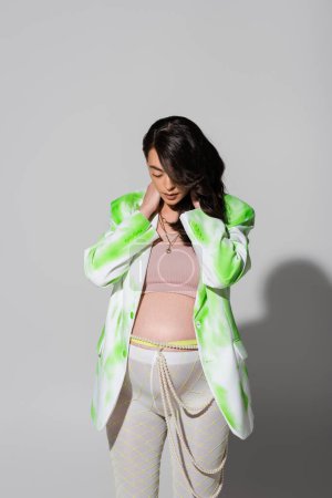 brunette pregnant woman in trendy green and white jacket, crop top, leggings and beads belt posing with hands on neck on grey background, stylish maternity concept, expectation