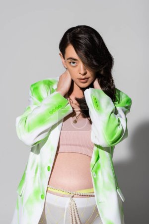 expecting mother with wavy brunette hair posing with hands on neck in green in white blazer, crop top, beads belt and leggings on grey background, maternity fashion concept, expectation