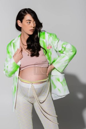 brunette pregnant woman in green and white blazer, crop top, beads belt and leggings holding hand on waist and looking away on grey background, maternity style concept, expectation