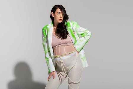 brunette pregnant woman in green and white jacket, tights, crop top, and beads belt standing with hand on hip on grey background, maternity style concept, expectation puzzle 656081556