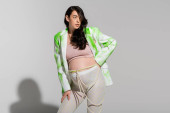 brunette pregnant woman in green and white jacket, tights, crop top, and beads belt standing with hand on hip on grey background, maternity style concept, expectation puzzle #656081556