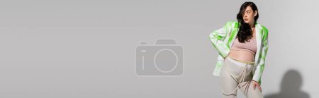 brünette schwangere Frau in grün-weißem Blazer, bauchfreiem Oberteil, Leggings und Perlengürtel stehend mit Hand auf Hüfte auf grauem Hintergrund, Erwartung, Umstandsmodekonzept, Banner