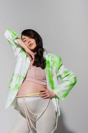 Schwangere mit welligem brünetten Haar posiert in bauchfreiem Top, grün-weißem Blazer, Perlengürtel und Leggings vor der Kamera auf grauem Hintergrund, modisches Umstandskonzept, Erwartung