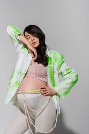 pregnant woman with wavy brunette hair, wearing green and white jacket, crop top, leggings and beads belt standing with hand on hip and looking away on grey background, trendy pregnancy concept