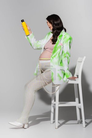volle Länge der schwangeren Frau mit glänzenden brünetten Haaren, trägt stylische Jacke, Leggings und Perlengürtel, sitzt auf einem Stuhl und hält Haarspray auf grauem Hintergrund, Umstandsmodekonzept