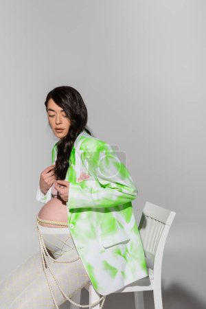 Schwangere mit welligem brünetten Haar, trendige Jacke, bauchfreies Oberteil, Perlengürtel und Leggings, auf Stuhl auf grauem Hintergrund sitzend, Umstandsmodekonzept, Erwartung