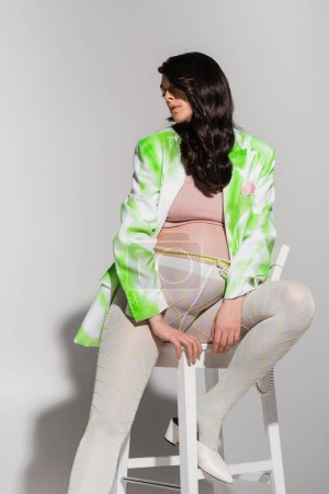 Téléchargez les photos : Femme enceinte aux cheveux bruns ondulés, vêtue d'un blazer vert et blanc, haut de culture, leggings et perles ceinture, posant sur chaise avec les yeux fermés sur fond gris, concept de mode maternité - en image libre de droit