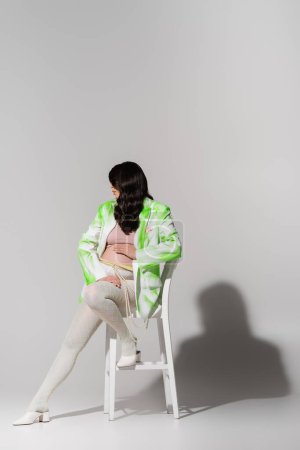 volle Länge der stilvollen werdenden Mutter mit welligen brünetten Haaren posiert auf Stuhl in grün-weißem Blazer, Crop Top, Perlengürtel und Leggings auf grauem Hintergrund, Umstandsmode-Konzept