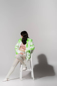 full length of stylish mom-to-be with wavy brunette hair posing on chair in green and white blazer, crop top, beads belt and leggings on grey background, maternity fashion concept puzzle #656081876