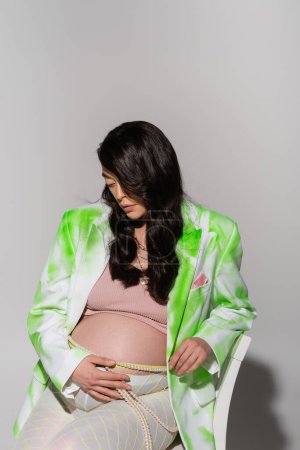 future mère avec des cheveux bruns ondulés touchant le ventre tout en posant en veston vert et blanc, crop top et ceinture de perles sur fond gris, concept de mode maternité, attente, femme enceinte 