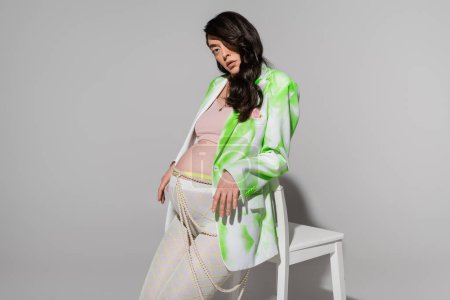 brunette mother-to-be in green and white blazer, leggings, crop top and beads belt looking at camera near chair on grey background, maternity fashion concept, expectation
