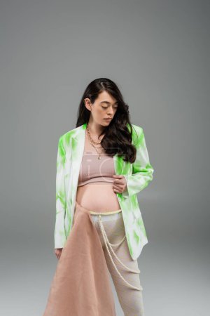brunette pregnant woman in green and white blazer, crop top, beads belt and leggings with beige chiffon cloth on grey background, maternity fashion concept