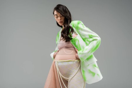 low angle view of brunette mother-to-be in green and white blazer, crop top, leggings with beige chiffon cloth and beads belt isolated on grey background, trendy pregnancy concept, expectation