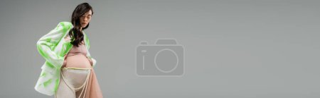 Tiefansicht der zukünftigen Mutter im trendigen grün-weißen Blazer, bauchfreies Oberteil, Leggings mit Perlengürtel und Chiffontuch isoliert auf grauem Hintergrund, Konzept Schwangerschaftsmode, Banner