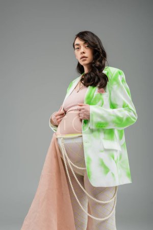 low angle view of charming mother-to-be in green and white blazer, crop top, beads belt and leggings with chiffon cloth looking at camera isolated on grey background, fashionable maternity concept