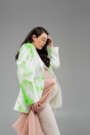 Téléchargez les photos : Vue latérale de la femme enceinte avec des cheveux bruns ondulés posant dans une veste élégante verte et blanche, leggings avec ceinture de perles et chiffon beige isolé sur fond gris, concept de mode maternité - en image libre de droit
