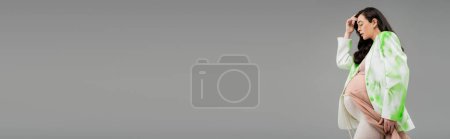 Téléchargez les photos : Vue latérale de la femme enceinte avec des cheveux bruns ondulés posant en veston vert et blanc, haut de culture et leggings avec chiffon et ceinture de perles isolé sur fond gris, bannière - en image libre de droit