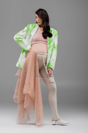 full length of brunette pregnant woman in green and white blazer, crop top and leggings with beige chiffon cloth and beads belt standing with hand on hip on grey background, maternity fashion concept