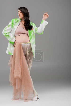 Brünette schwangere Frau in grün-weißer Jacke, bauchfreiem Oberteil, Perlengürtel und Leggings posieren mit beigem Chiffontuch auf grauem Hintergrund, Umstandsmodekonzept, Erwartung