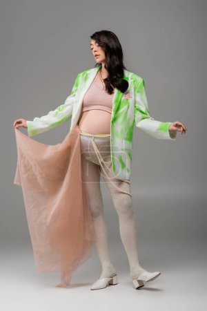 full length of charming mom-to-be in trendy jacket, crop top and leggings with beads belt posing with beige chiffon cloth on grey background, expectation, fashionable pregnancy concept