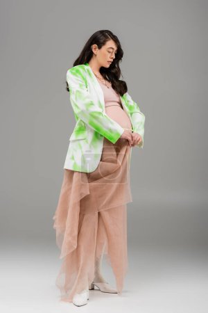 full length of pregnant woman with wavy brunette hair posing in green and white blazer, crop top and beige chiffon cloth on grey background, maternity fashion concept, expectation