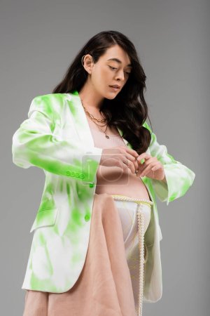 werdende Mutter in grün-weißer Jacke, bauchfreies Oberteil, Leggings mit Perlengürtel und Chiffontuch posieren mit bäuchennahen Händen isoliert auf grauem Hintergrund, modisches Umstandskonzept