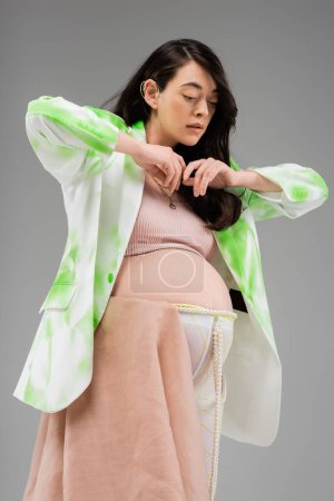 pregnant woman with wavy brunette hair, wearing crop top, green and white blazer, leggings with chiffon cloth and beads belt isolated on grey background, maternity style concept, expectation