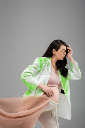 brunette pregnant woman in green and white blazer, crop top, beads belt and leggings with chiffon cloth isolated on grey background, maternity fashion concept, expectation