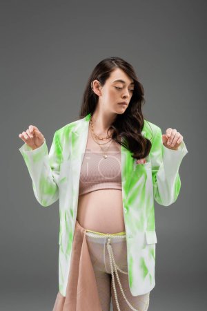 Ansprechende werdende Mama im bauchfreien Top, trendiger grün-weißer Blazer, Perlengürtel und Strumpfhose mit beigem Chiffontuch auf grauem Hintergrund, modisches Schwangerschaftskonzept, Erwartung