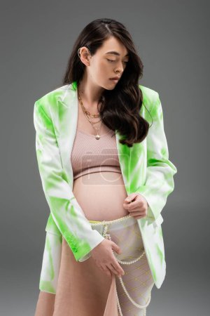 expecting mother in green and white blazer, crop top, leggings with chiffon cloth and beads belt standing isolated on grey background, maternity fashion concept, pregnant woman 
