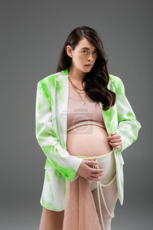 expecting mother with wavy brunette hair, wearing green blazer, crop top and leggings with beads belt and chiffon cloth, looking at camera isolated on grey background, fashionable maternity concept