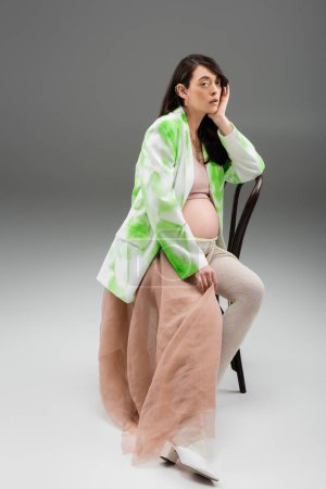 full length of appealing pregnant woman in stylish jacket, crop top and leggings with beads belt and beige chiffon sitting on chair and looking at camera on grey background, maternity fashion concept