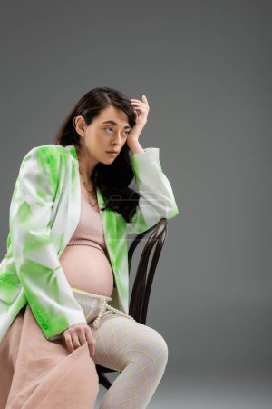 charming brunette mother-to-be in fashionable blazer, crop top and leggings with beads belt and chiffon cloth sitting on chair and looking away on grey background, maternity style concept
