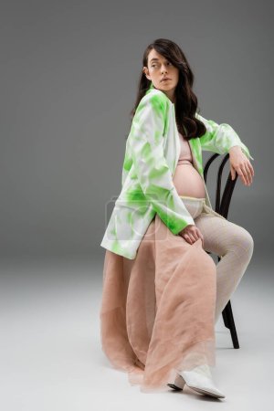 full length of brunette mother-to-be in stylish jacket and leggings with beads belt and beige chiffon cloth sitting on chair and looking away on grey background, fashionable pregnancy concept
