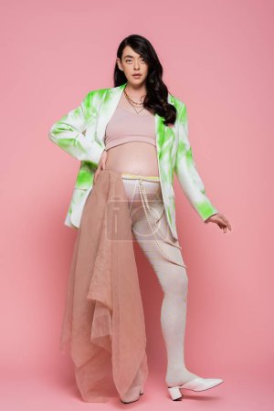 full length of expecting mother in green and white jacket, crop top, beads belt and leggings with chiffon cloth standing with hand on hip on pink background, maternity fashion concept