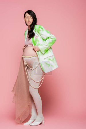 full length of pregnant woman with wavy brunette hair posing in fashionable blazer, beads belt and leggings with beige chiffon cloth on pink background, maternity fashion concept