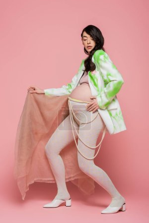 Brünette schwangere Frau in trendigem Blazer, bauchfreiem Oberteil, Perlengürtel und Leggins posiert mit beigem Chiffontuch auf rosa Hintergrund, Umstandsmodekonzept