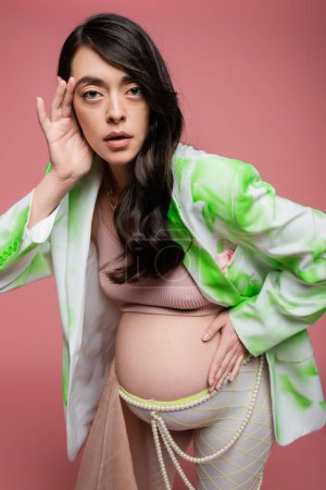 brunette mom-to-be wearing crop top with green and white blazer, holding hand near face while looking at camera isolated on pink, maternity fashion concept, pregnant woman 