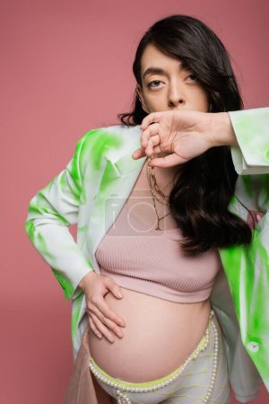 brunette pregnant woman in crop top, fashionable blazer and beads belt holding hand near face, touching tummy and looking at camera isolated on pink background, maternity style concept