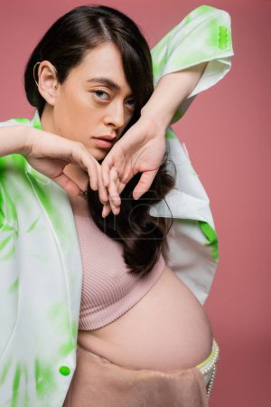 fashionable pregnant woman in crop top with green and white blazer holding hands near face and looking at camera isolated on pink background, maternity style concept
