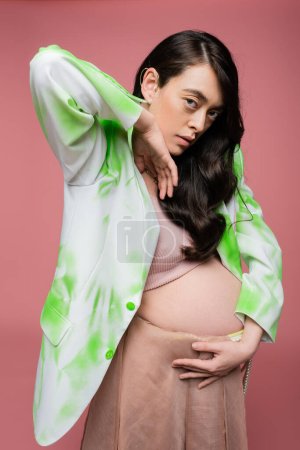 brunette mother-to-be holding hand near face and looking at camera while posing in green and white blazer and crop top isolated on pink background, maternity fashion concept