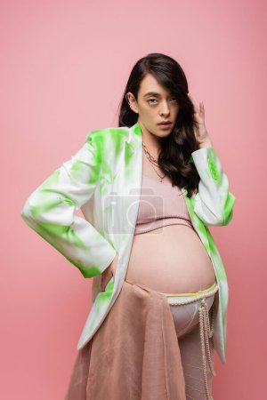 appealing future mother in green and white blazer, crop top and leggings with beads belt and chiffon cloth looking at camera isolated on pink background, stylish pregnancy concept