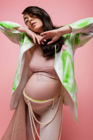 brunette pregnant woman in green and white blazer, crop top, leggings with beads belt and chiffon cloth holding hands under chin and looking at camera isolated on pink, maternity fashion concept