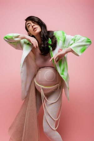 charming mom-to-be posing in green and white jacket, crop top, leggings with beads belt and beige chiffon cloth isolated on pink background, trendy maternity concept, pregnant woman 