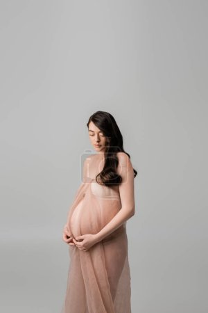 romántica madre-a-ser con cabello castaño ondulado y pulseras doradas de pie en tela de gasa beige y suave aislado sobre fondo gris, concepto de moda de maternidad, mujer embarazada 