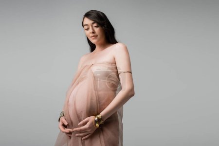 femme enceinte heureuse et élégante en mousseline de soie beige et bracelets dorés debout et souriants isolés sur fond gris, concept de mode maternité, sensualité 