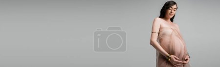 Foto de Mujer embarazada romántica y elegante en gasa aireada drapeado y accesorios de oro de pie aislado sobre fondo gris, concepto de moda de maternidad, bandera, sensualidad - Imagen libre de derechos